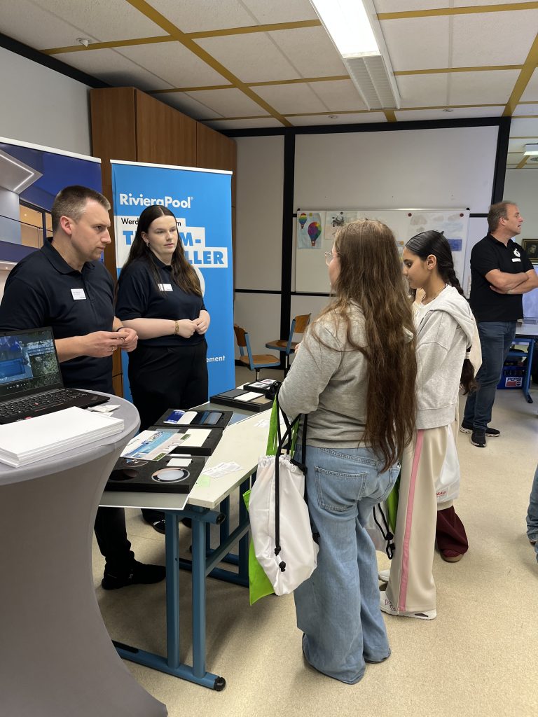 Zwei Jugendliche sprechen mit Ausstellern an einem Messestand im Klassenraum.